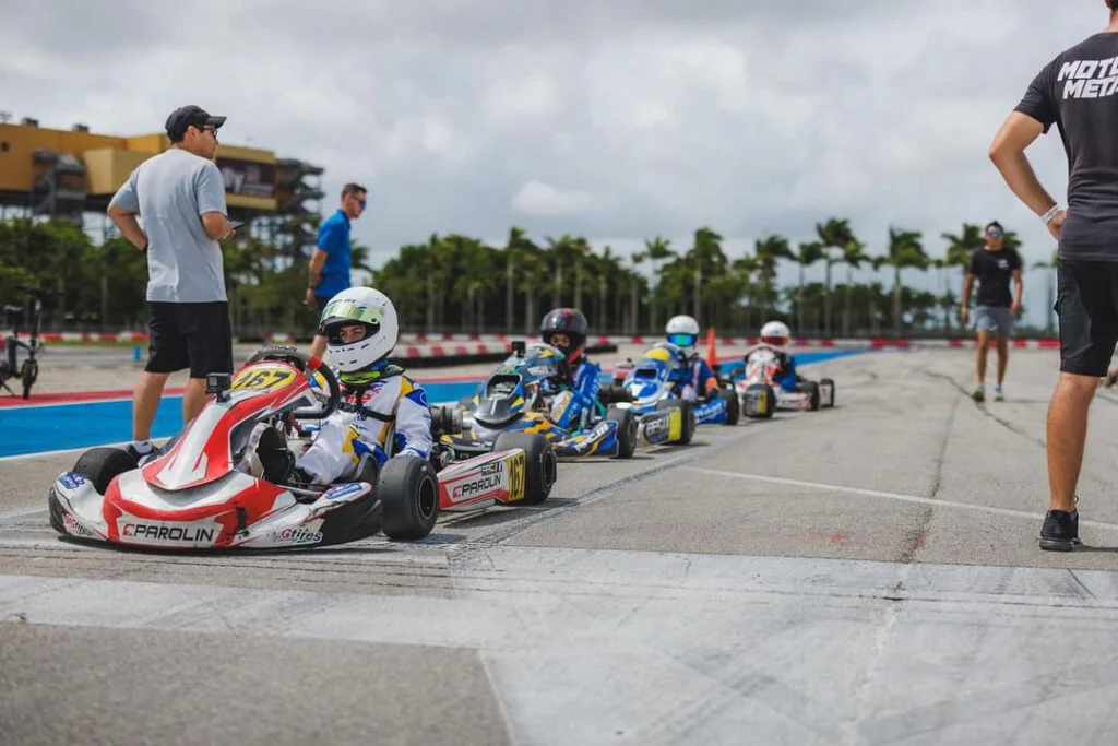 4 High speed go kart racers lined up on a track in their karts with 2 trainers standing by, instructing